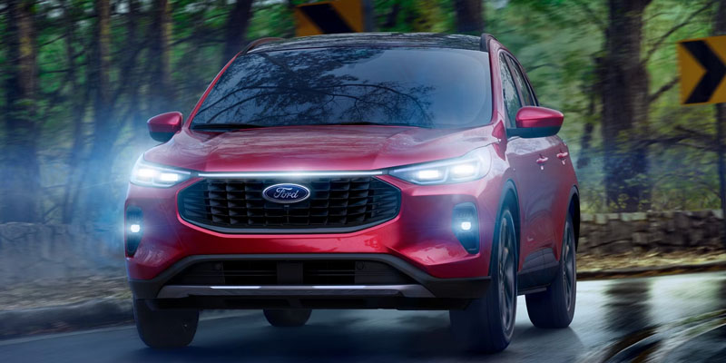Front exterior view of a red 2026 Ford Escape driving on a forest road, featuring LED headlights, black grille, and modern compact SUV styling.
